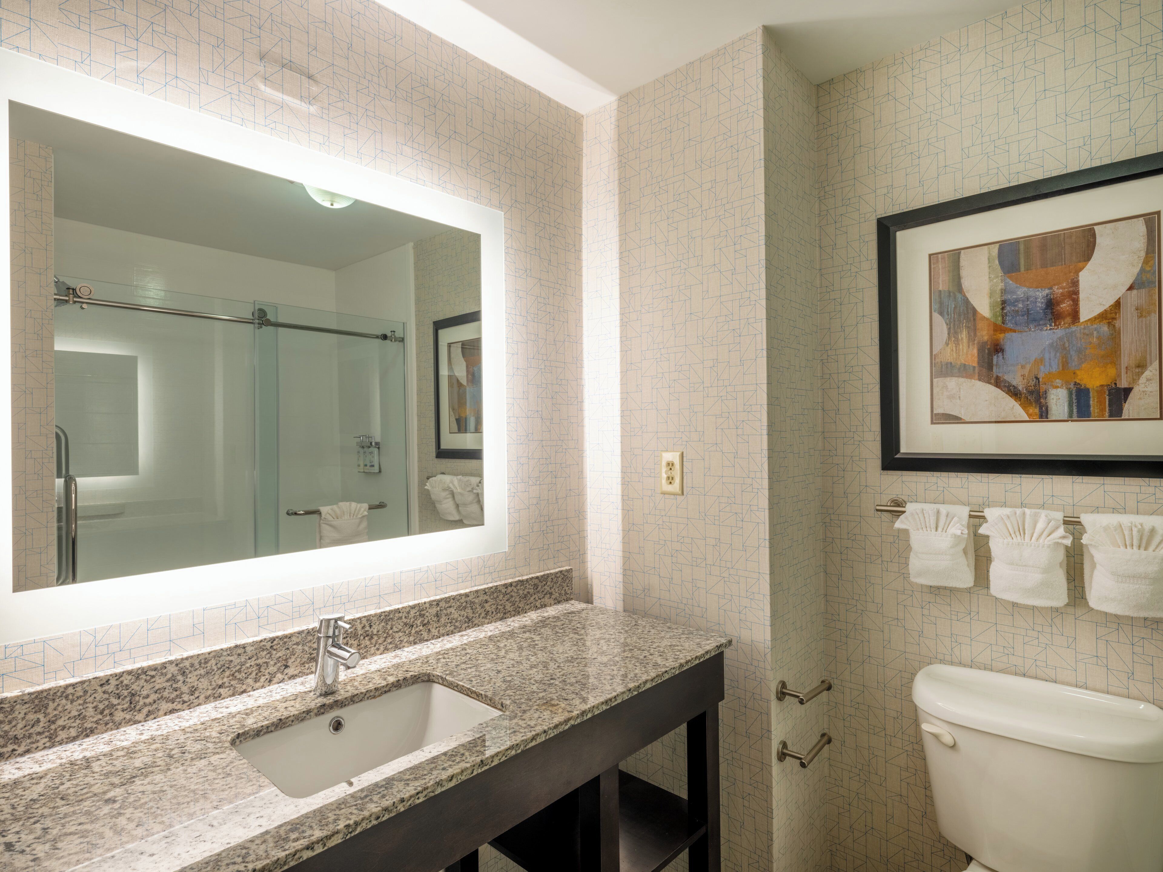 A bathroom with a granite countertop sink, a large mirror, a framed abstract painting, and a toilet with three towels hanging on the wall.