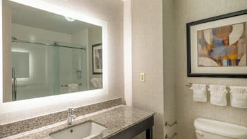 A bathroom with a granite countertop sink, a large mirror, a framed abstract painting, and a toilet with three towels hanging on the wall.