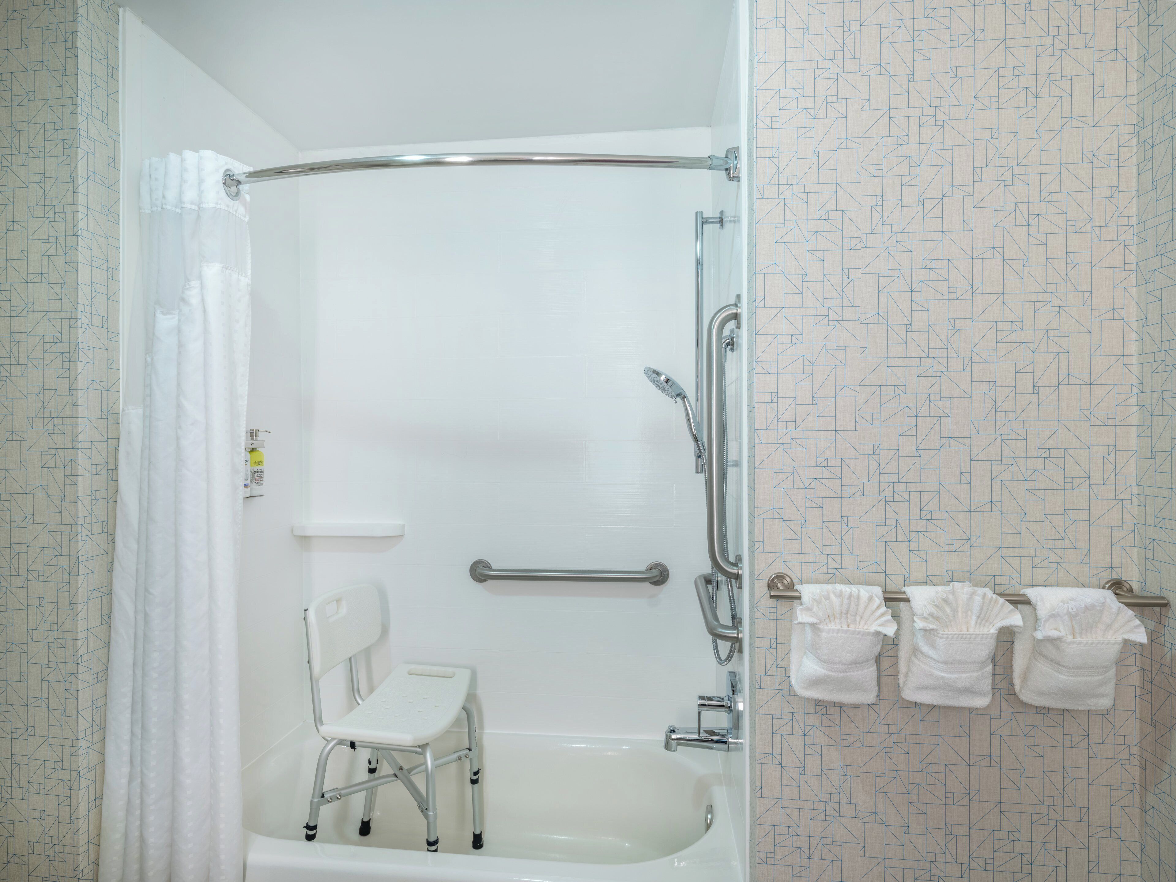 A bathroom with a shower, a white bathtub, a white chair, and a towel rack with towels.