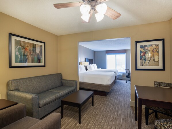 Ein Hotelzimmer mit grauem Sofa, einem kleinen Tisch, einem Bett mit weißer Bettwäsche, einem Deckenventilator und zwei gerahmten Kunstwerken an den Wänden.