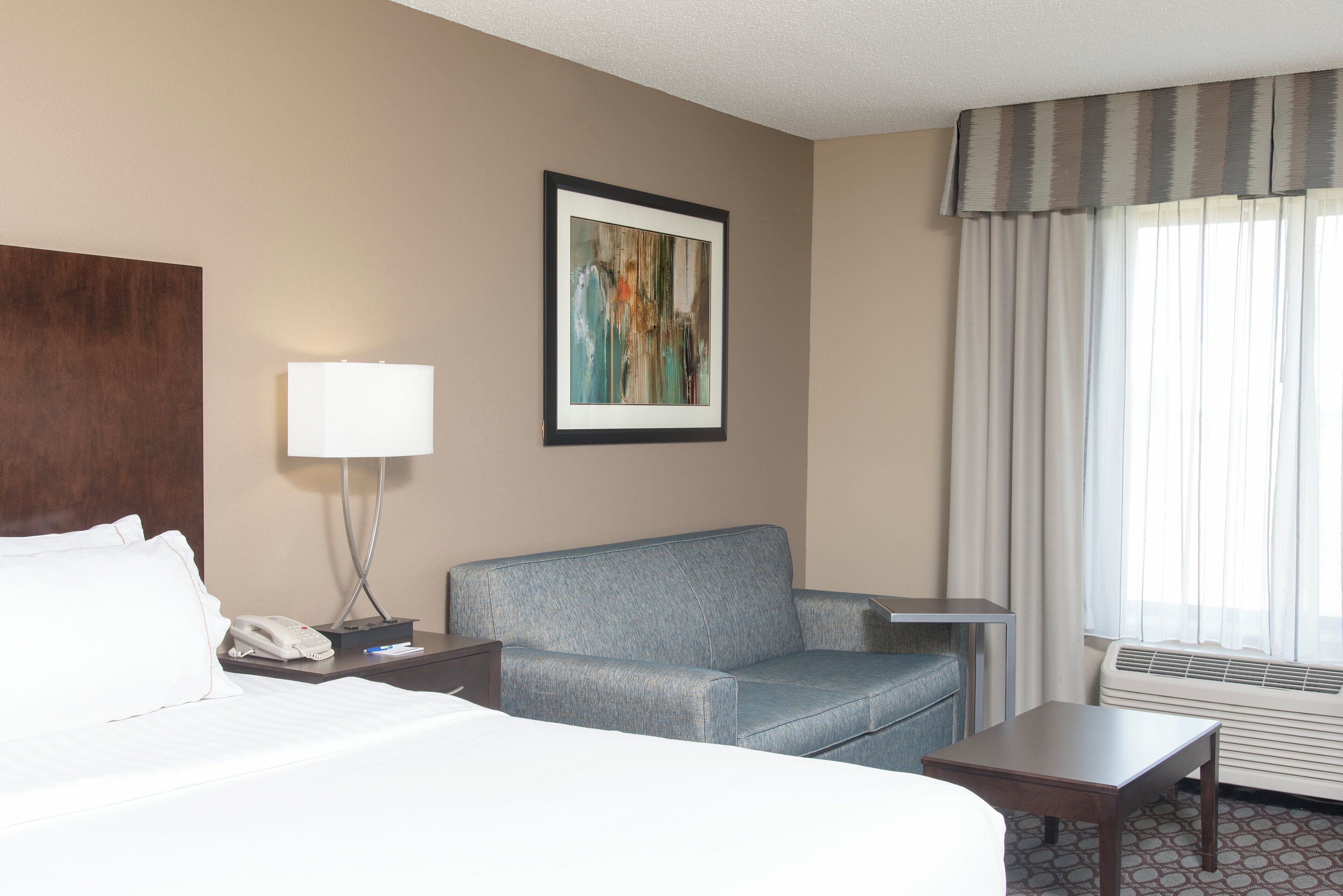 A hotel room with a bed, a desk, a chair, a lamp, a phone, a painting, and a window with curtains.