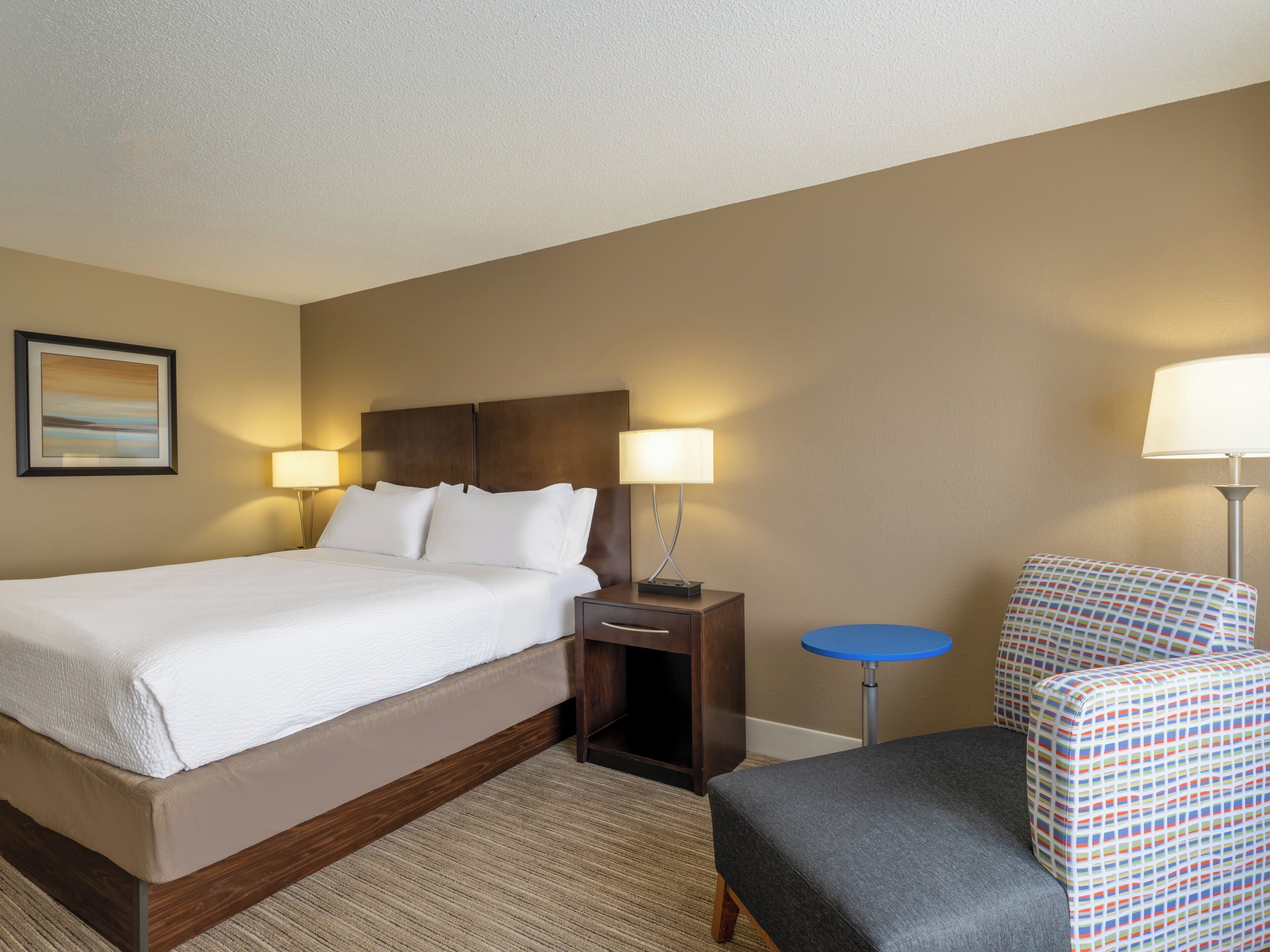 A hotel room with a large bed, two bedside lamps, a small round table, a patterned armchair, and a framed picture on the wall.