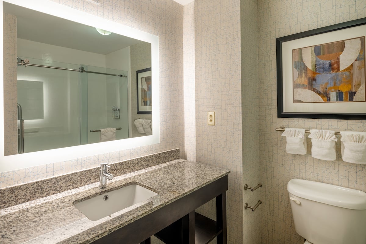 A bathroom with a granite countertop sink, a large mirror, a framed abstract painting, and a toilet with three towels hanging on the wall.