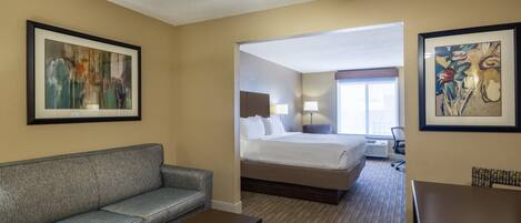 A hotel room with a bed, a sofa, a coffee table, and a dining table. There is a ceiling fan, a window, and artwork on the walls.