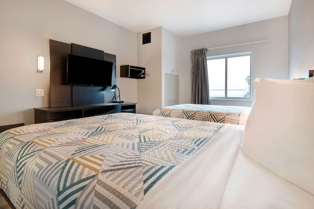 A hotel room with two beds, a flat-screen TV, a desk, and a window with curtains.