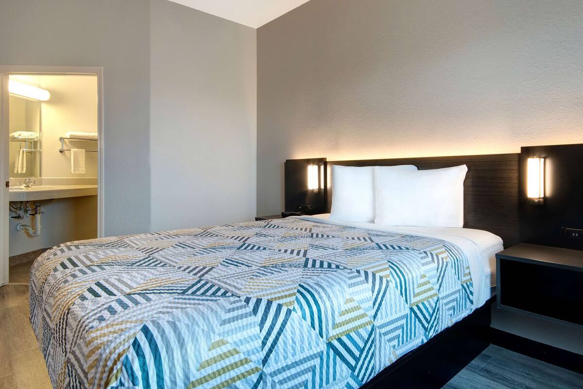 A hotel room with a large bed, a nightstand, a wall-mounted lamp, and a bathroom visible through an open door.