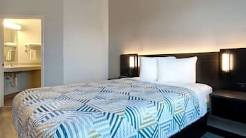 Une chambre d’hôtel avec un grand lit, une table de chevet, une lampe fixée au mur et une salle de bain visible à travers une porte ouverte.