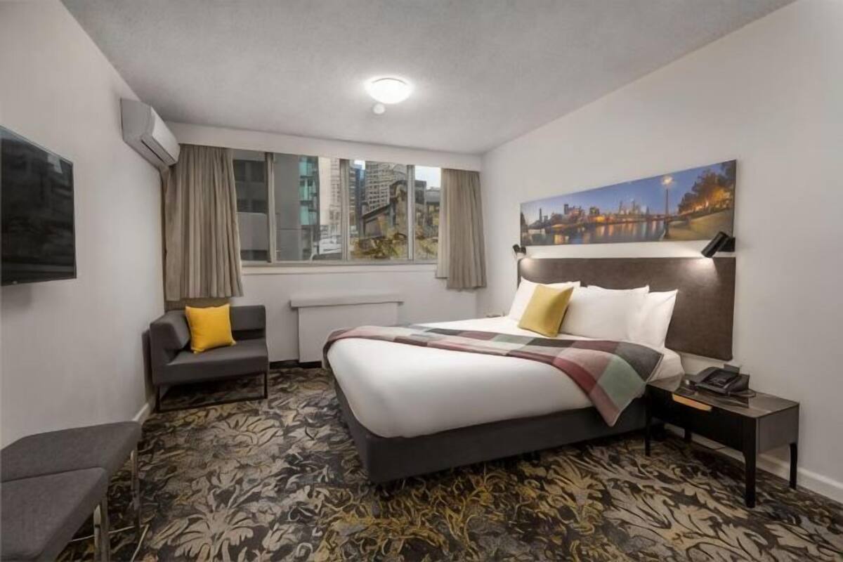 Habitación de hotel con cama, escritorio, silla, ventana con cortinas y un cuadro en la pared.