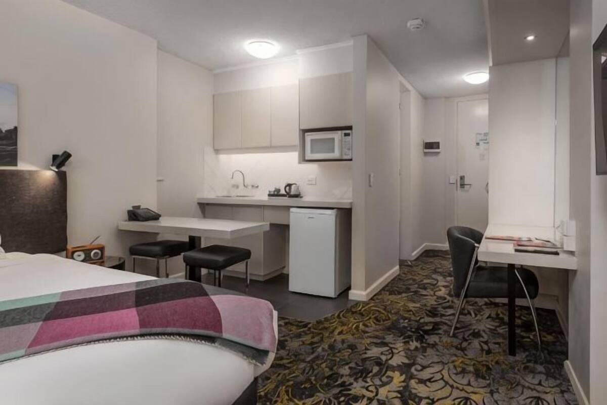 Habitación de hotel con cama, escritorio, silla, microondas, kitchenette pequeña y alfombra estampada.