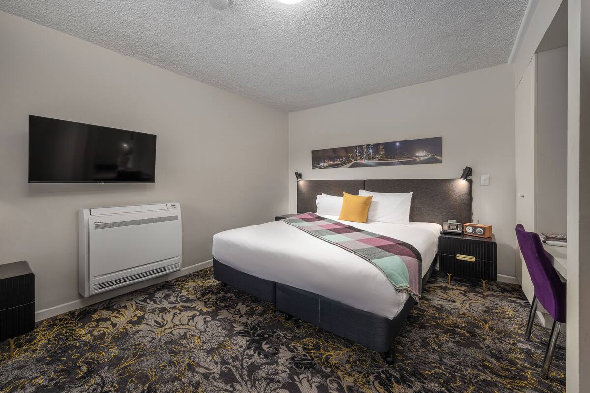 Habitación de hotel con cama, televisor de pantalla plana montado en la pared, calefactor, mesita de noche con radio y alfombra estampada.