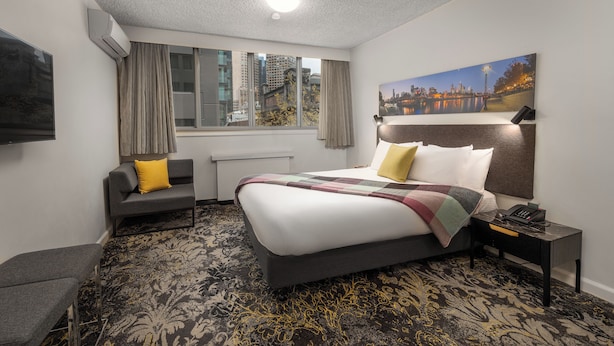 Habitación de hotel con una cama grande, una mesita de noche con lámpara, una silla, una ventana con cortinas y vistas a la ciudad.