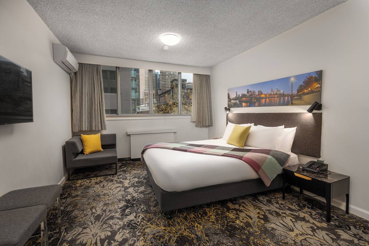 Habitación de hotel con una cama grande, una mesita de noche con lámpara, una silla, una ventana con cortinas y vistas a la ciudad.