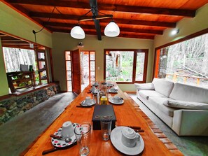 Un rincón de comedor con una mesa de madera dispuesta para el té, un sofá blanco y grandes ventanales con vista al bosque.