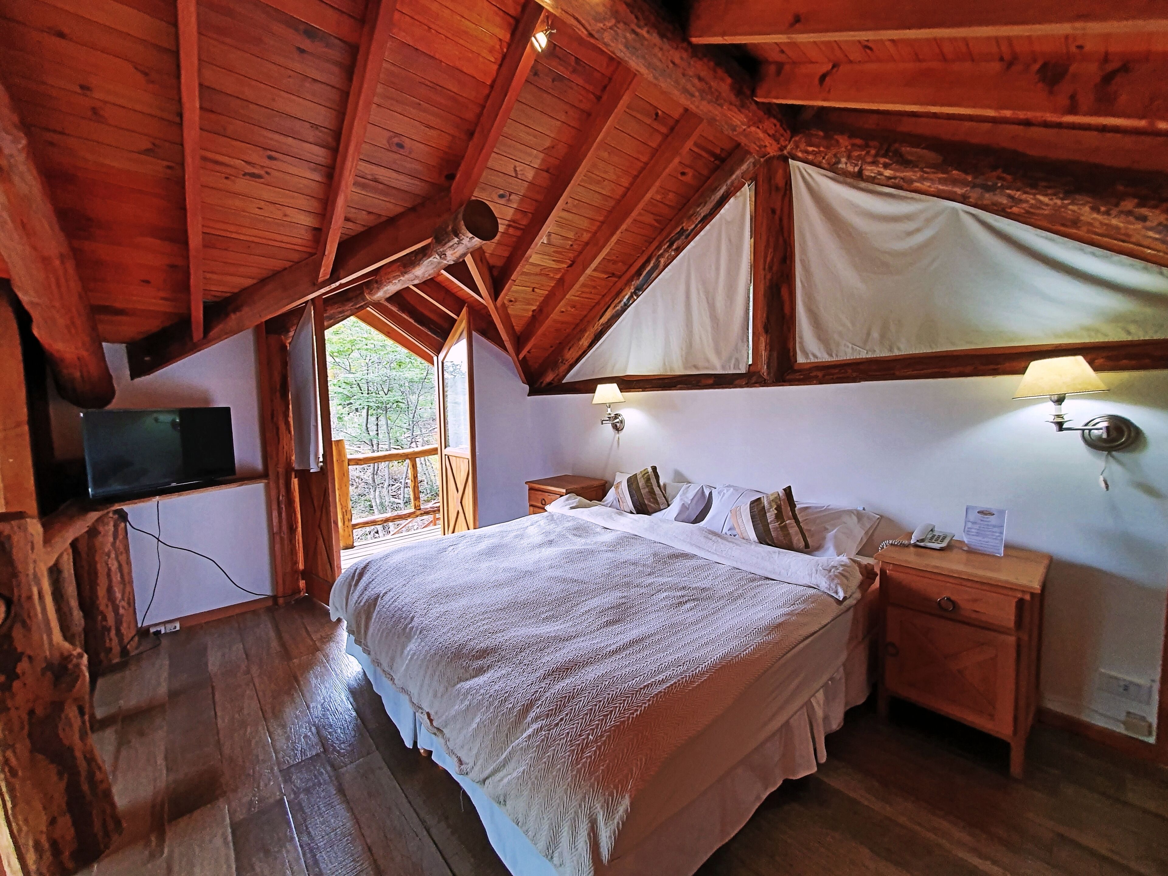Un dormitorio de cabaña de madera con cama, lámparas de noche, una mesita de noche de madera y un televisor sobre un mueble de madera.