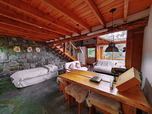 Interior rústico de una cabaña con pared de piedra, vigas de madera, una cama en altillo, una mesa de comedor y un sofá.