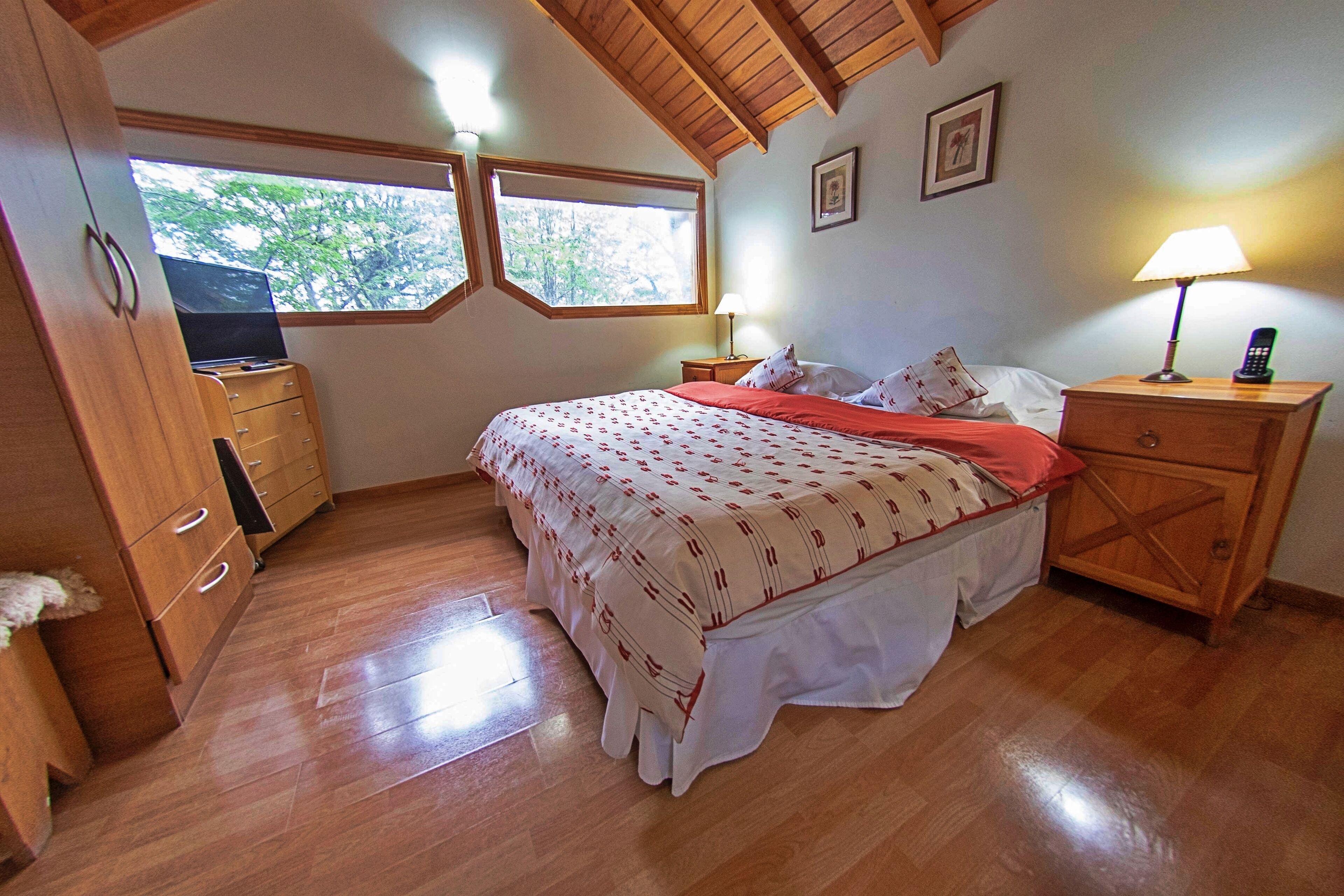 A bedroom with a bed, wooden dresser, nightstands, and a window with a view of trees.