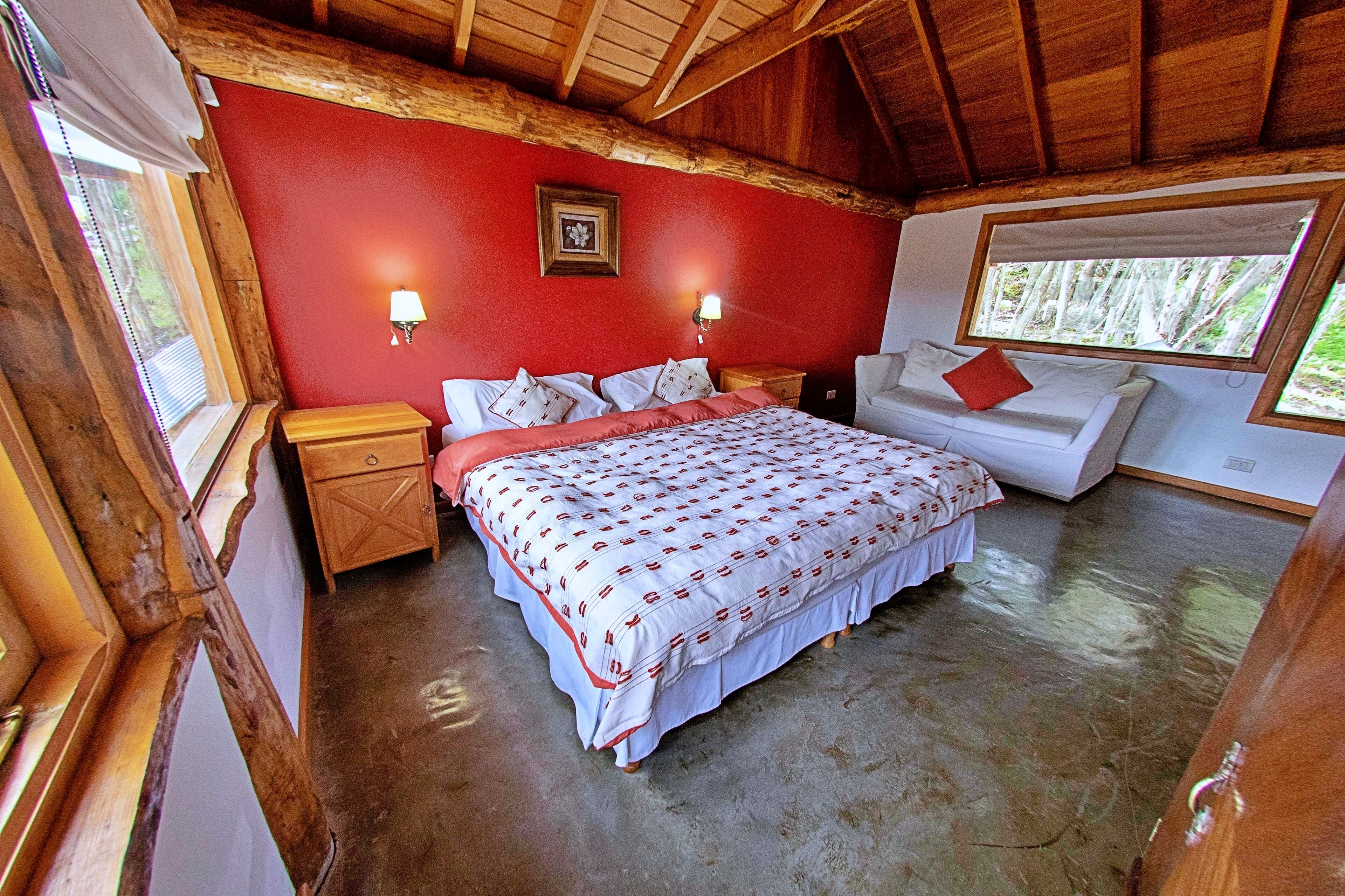A bedroom with a large bed, a nightstand, a dresser, a window with a view of trees, and a red wall.