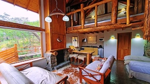 L’intérieur d’un chalet rustique, avec une cheminée en pierre, des meubles en bois, un coin cuisine et une vue sur la forêt environnante.