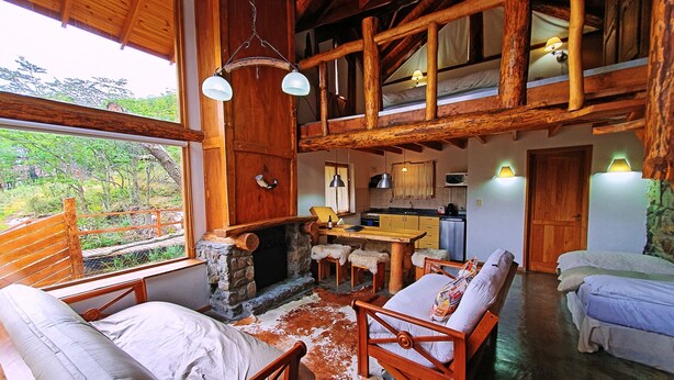 A rustic cabin interior with a stone fireplace, wooden furniture, a kitchen area, and a view of the surrounding forest. - Patagonia Villa (Ushuaia)