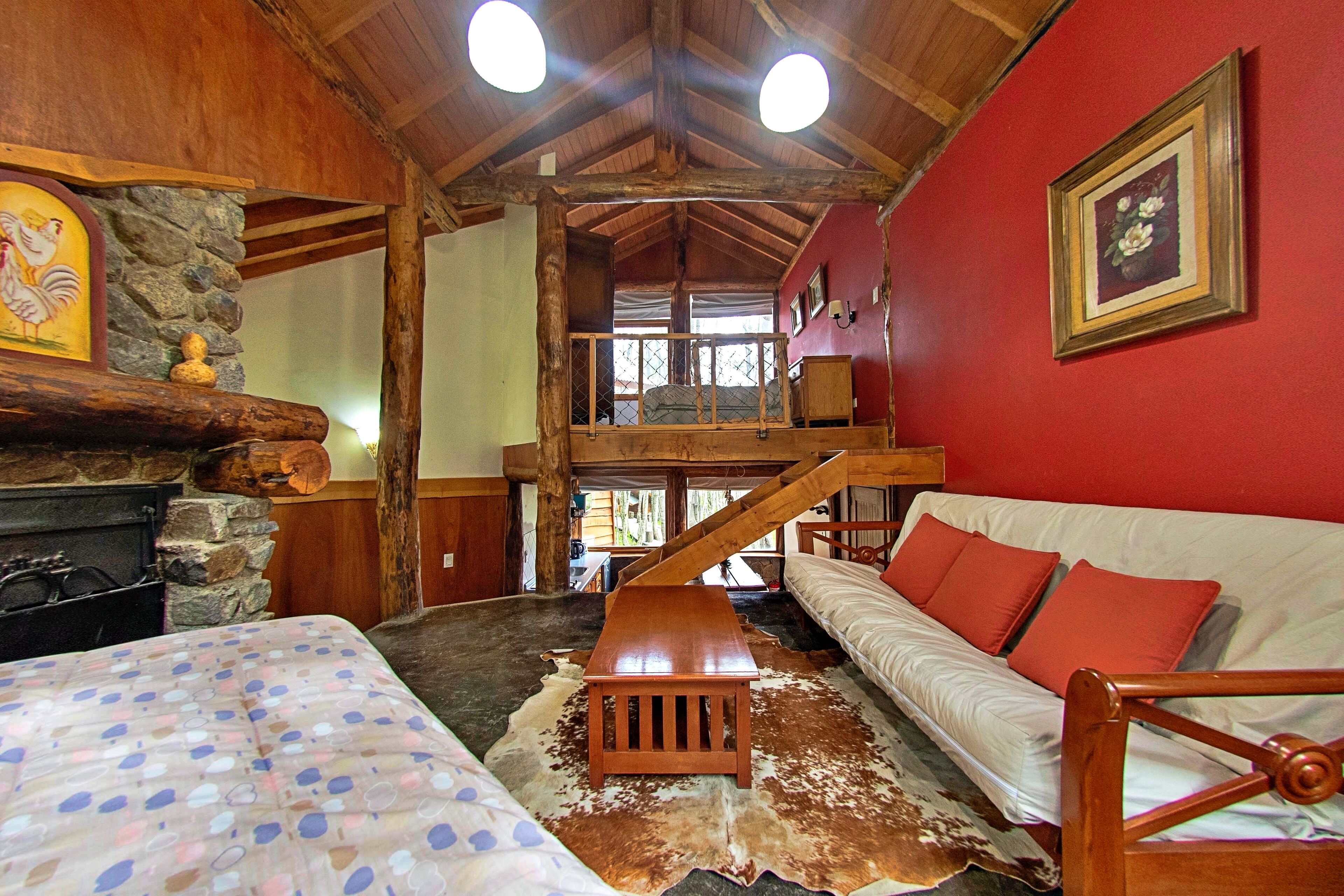 Interior rústico de una cabaña con techo de madera, chimenea de piedra, zona de altillo y habitación con paredes rojas, sofá y cojines.