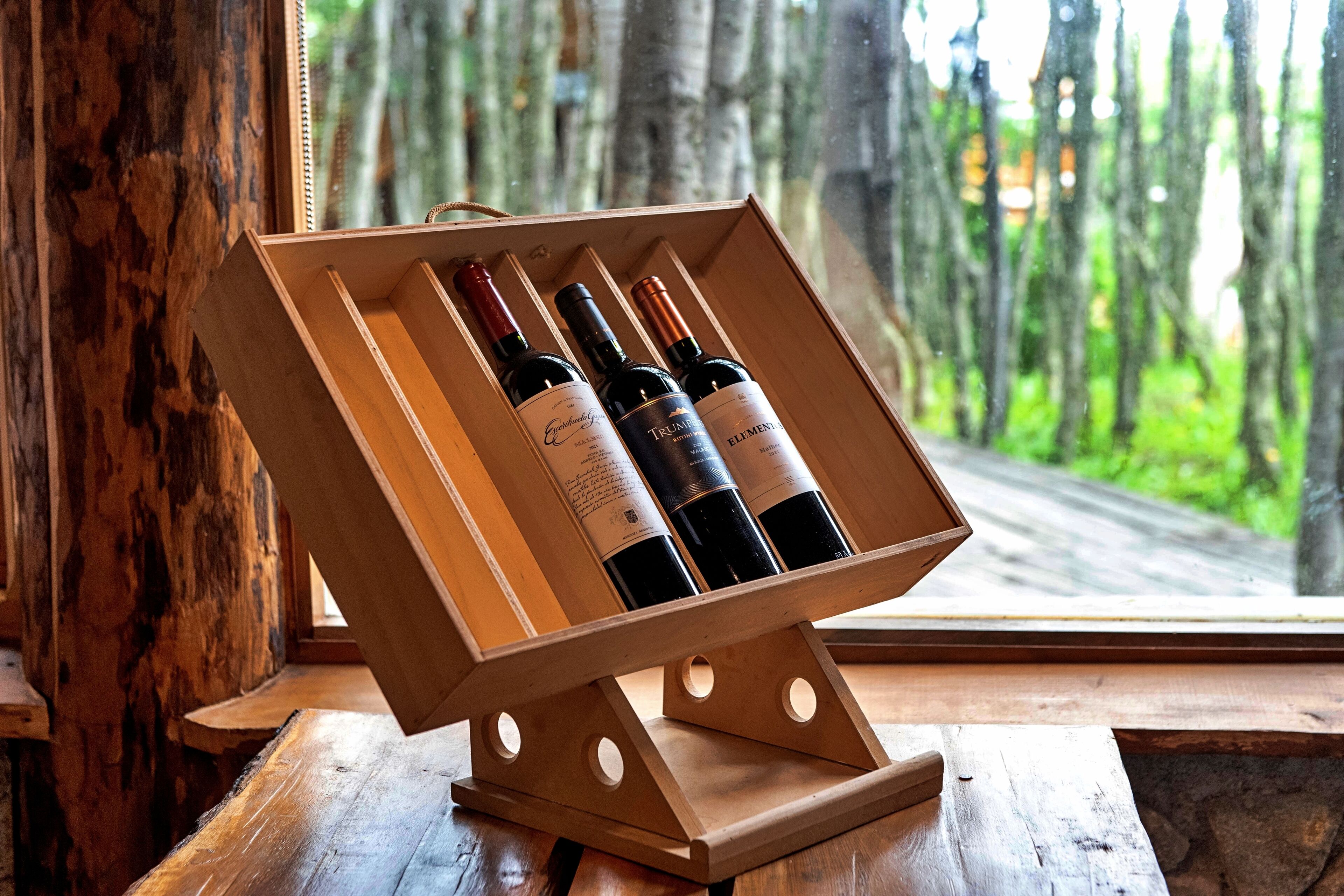 Een houten wijnrek met drie flessen wijn, voor een raam met uitzicht op een bos.