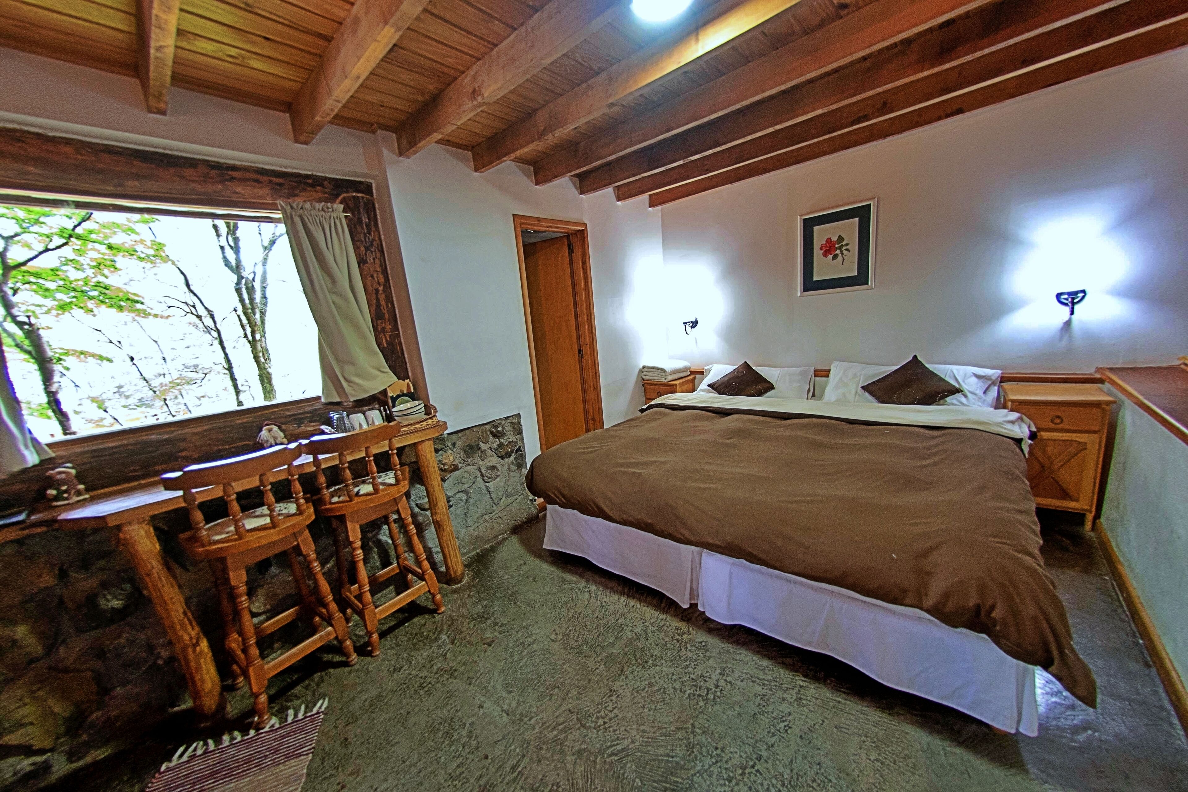 Un dormitorio con cama de madera, un escritorio con sillas, una pared de piedra y una ventana con cortinas.