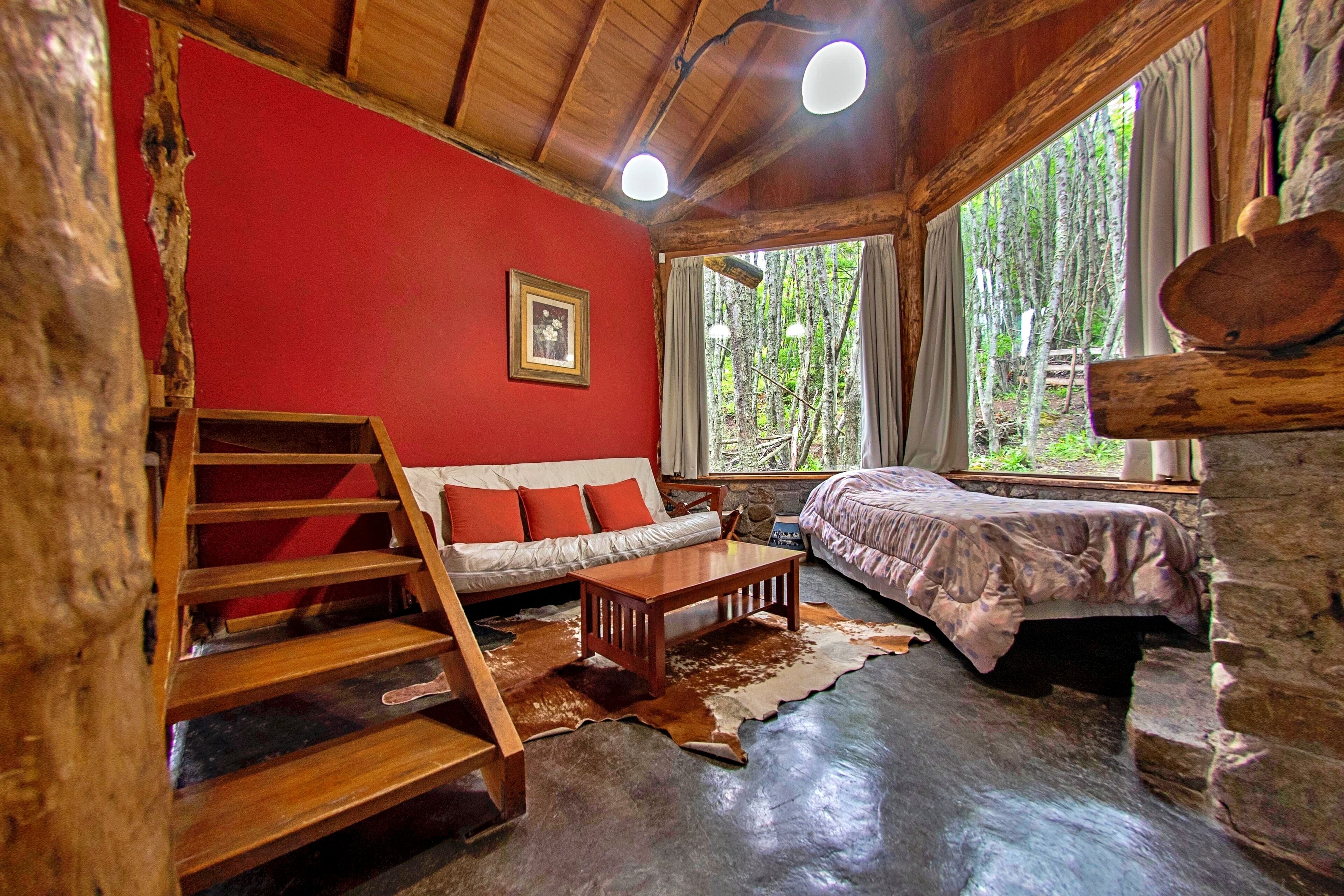 A rustic cabin interior with a wooden staircase, a sofa, a bed, and a large window with a view of trees.