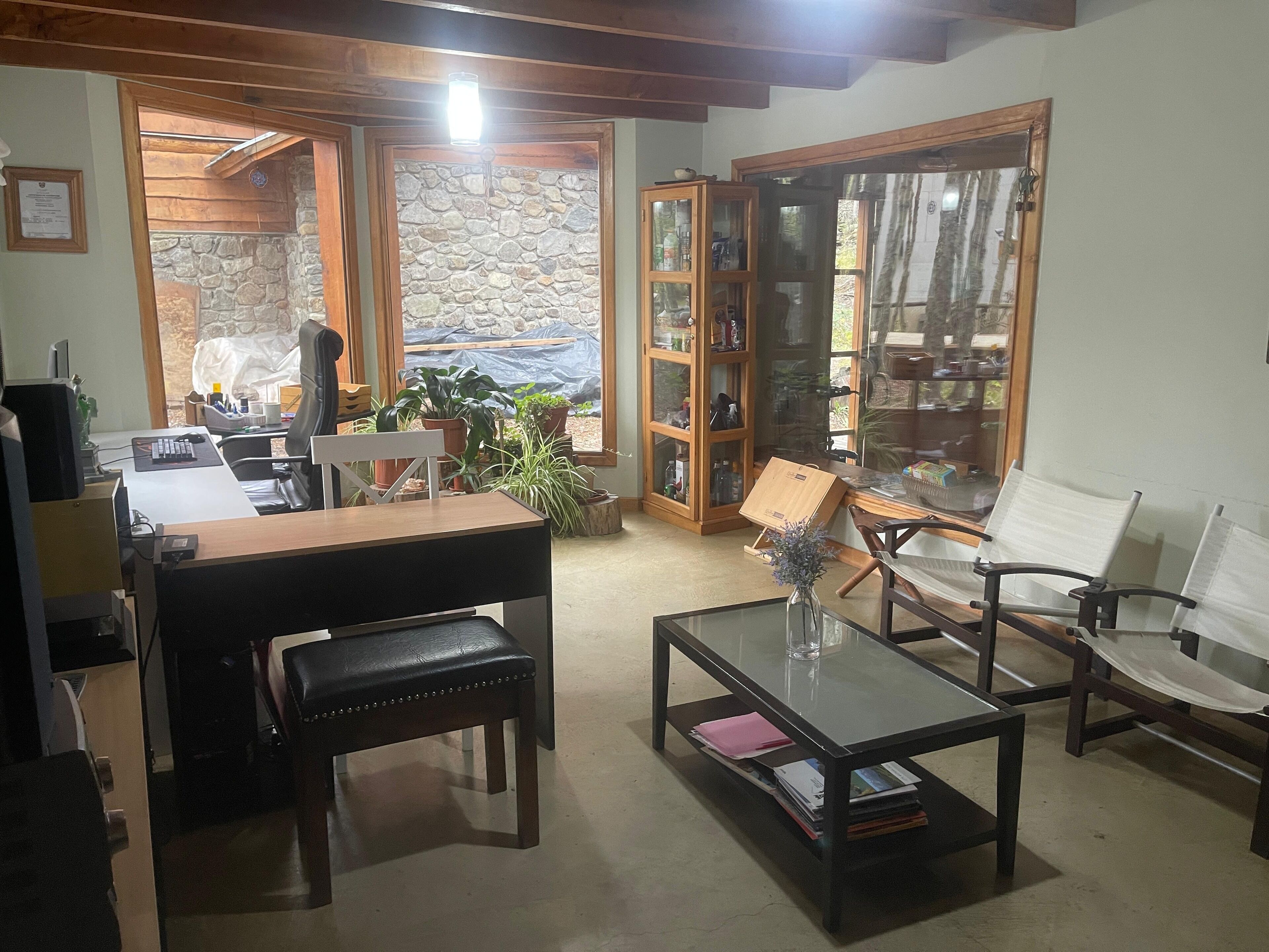 A cozy living room with a wooden desk, a glass coffee table, and comfortable seating. There is a stone wall and a large window with curtains.