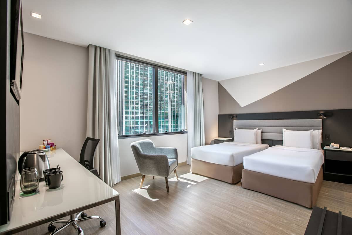Habitación de hotel con dos camas, un escritorio, una silla y un amplio ventanal con cortinas.