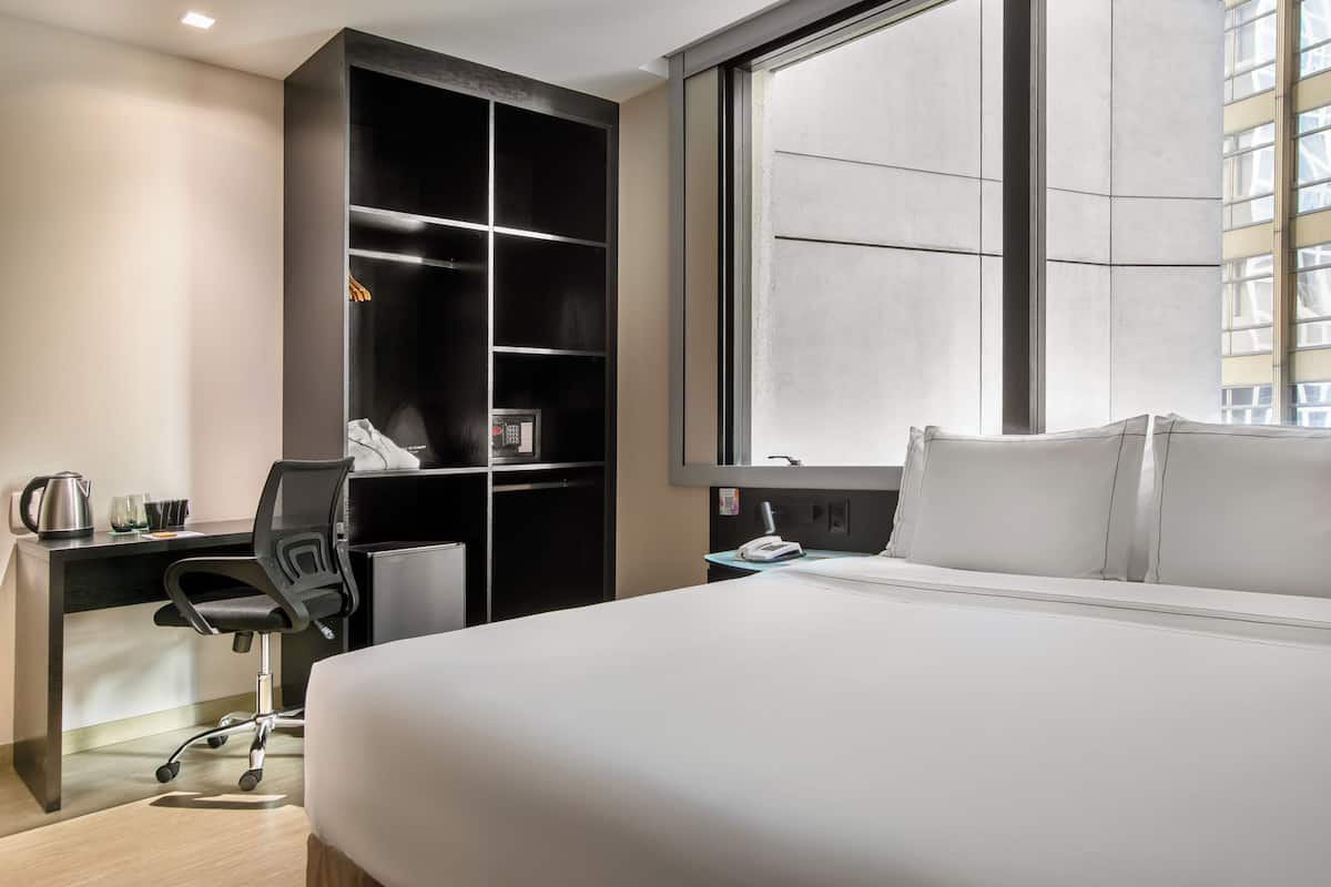Habitación de hotel con cama, escritorio, silla, armario y ventana grande.