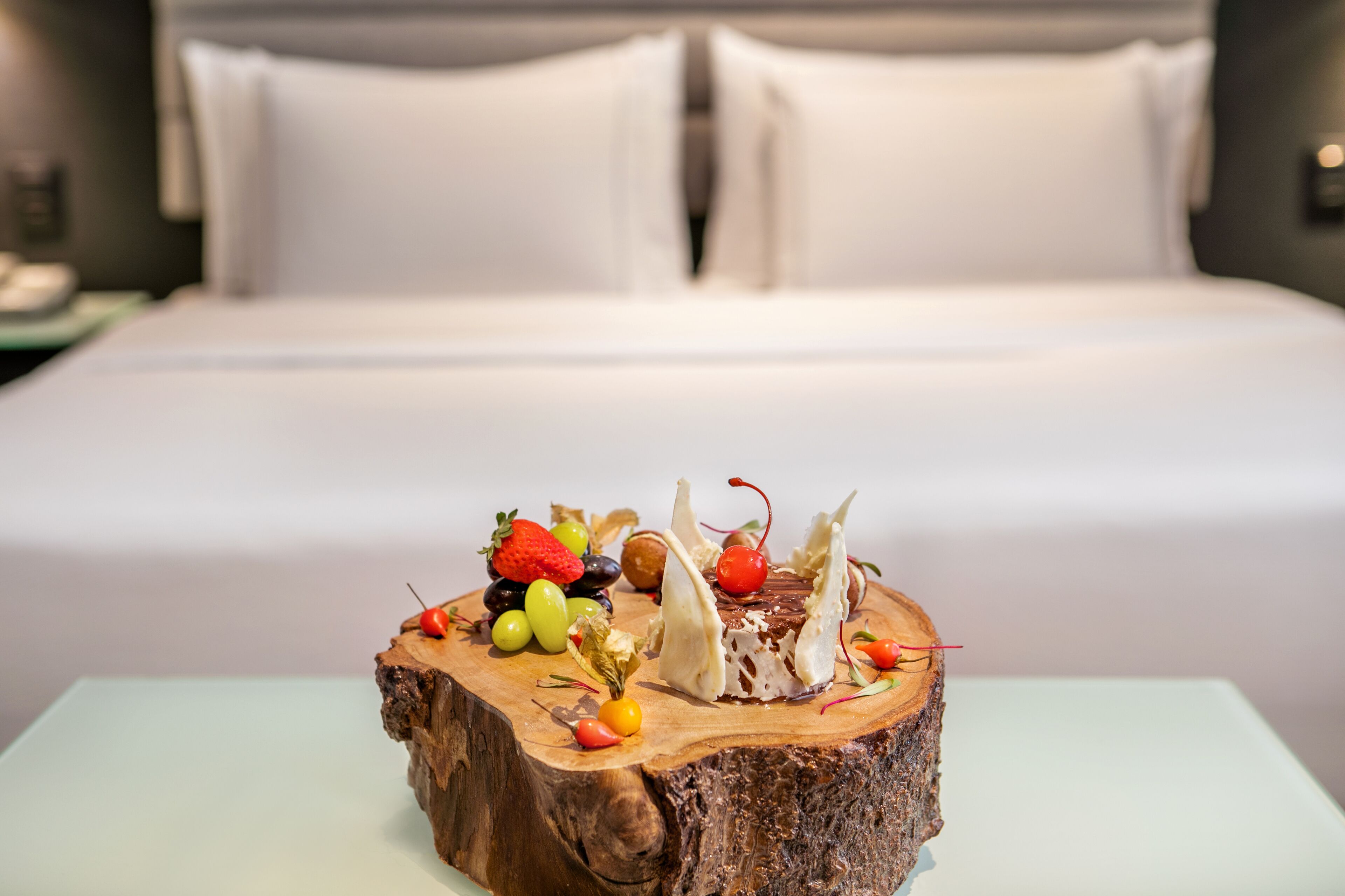 Una cama con sábanas y almohadas blancas, una mesita de noche de madera con una lámpara y un postre de fruta y chocolate sobre un tocón de árbol.