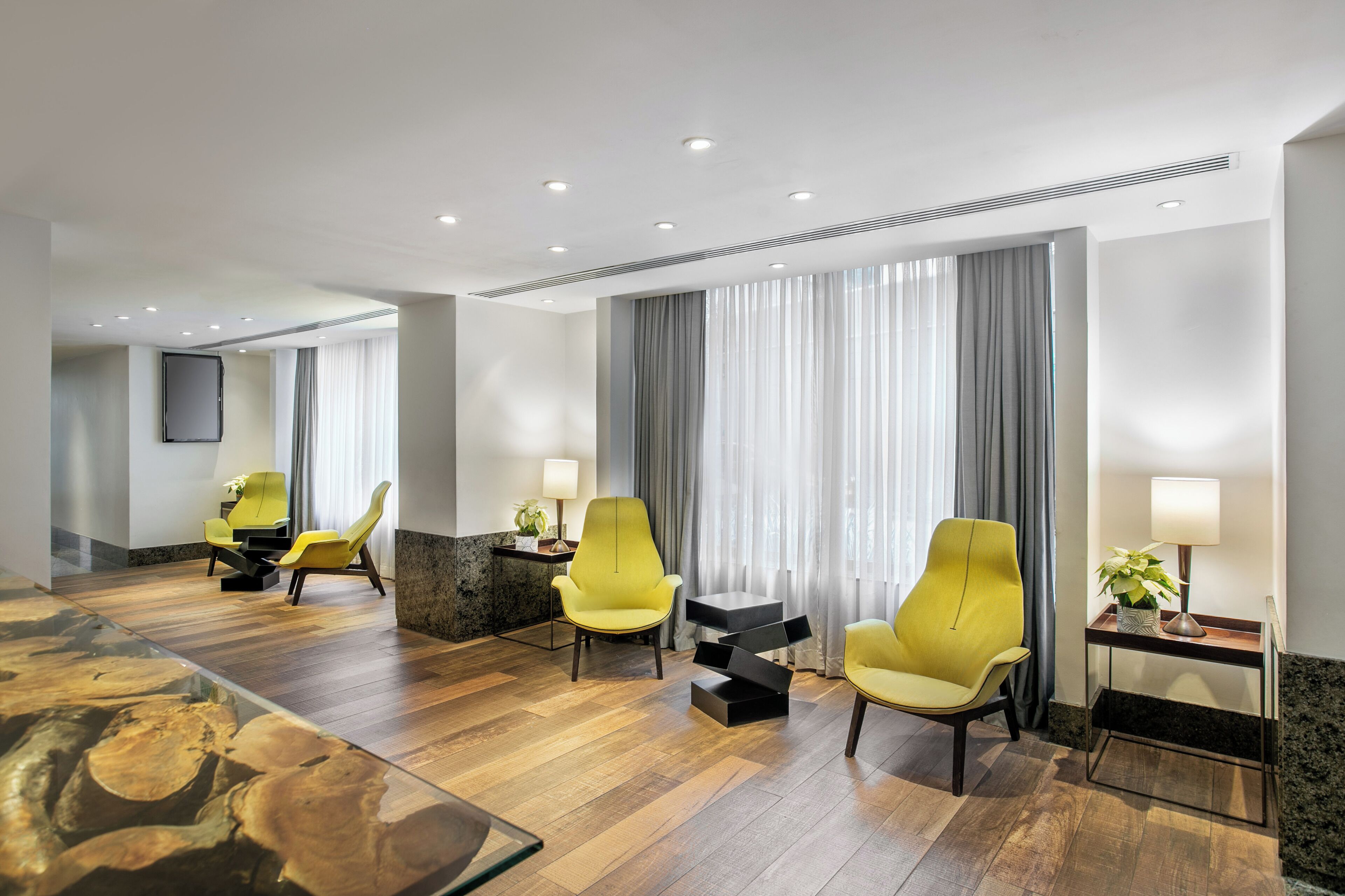 A modern hotel room with wooden flooring, yellow armchairs, a glass coffee table, and a large mirror.