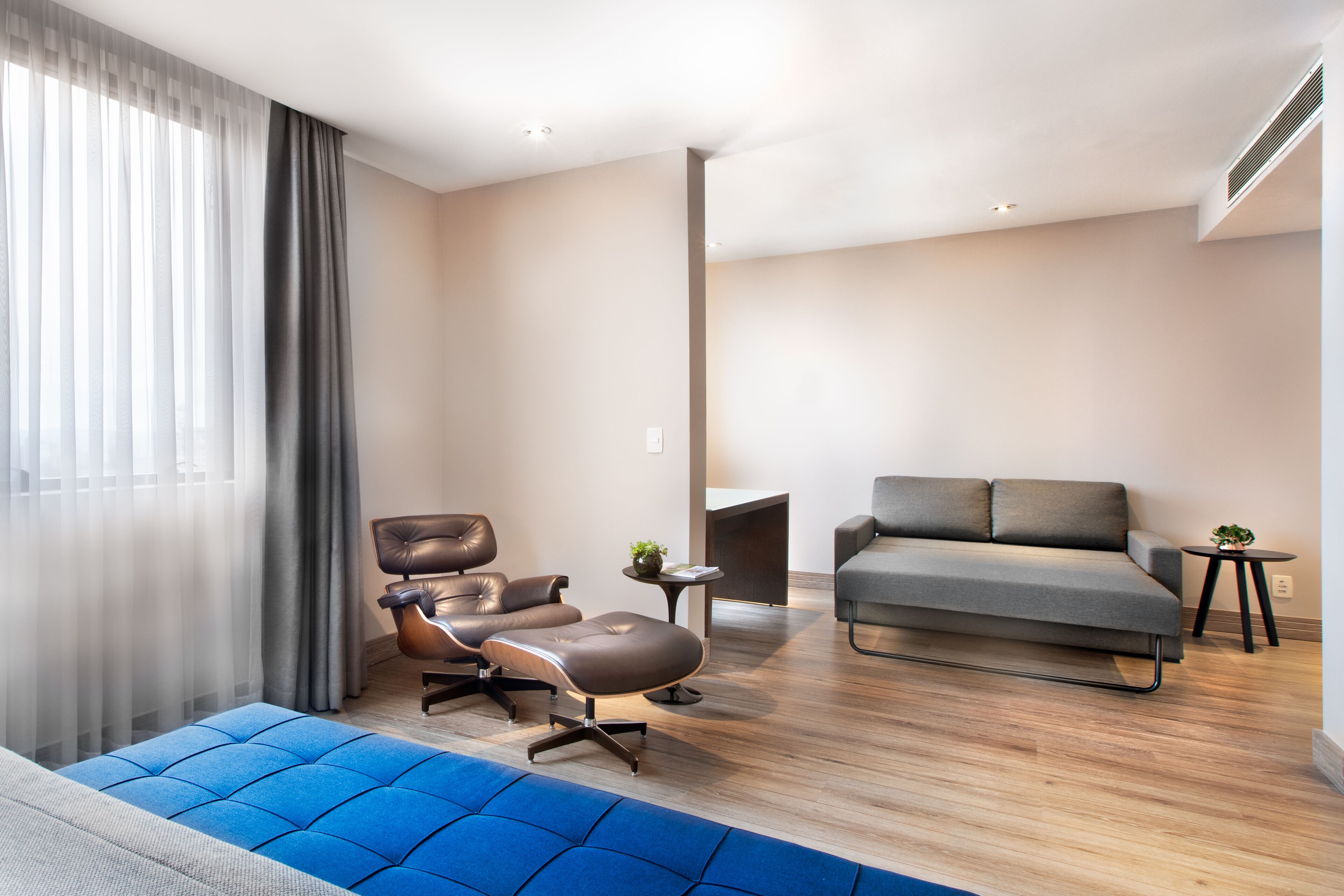 A modern living room with a sofa, a leather armchair, a blue ottoman, and a small table with a plant.