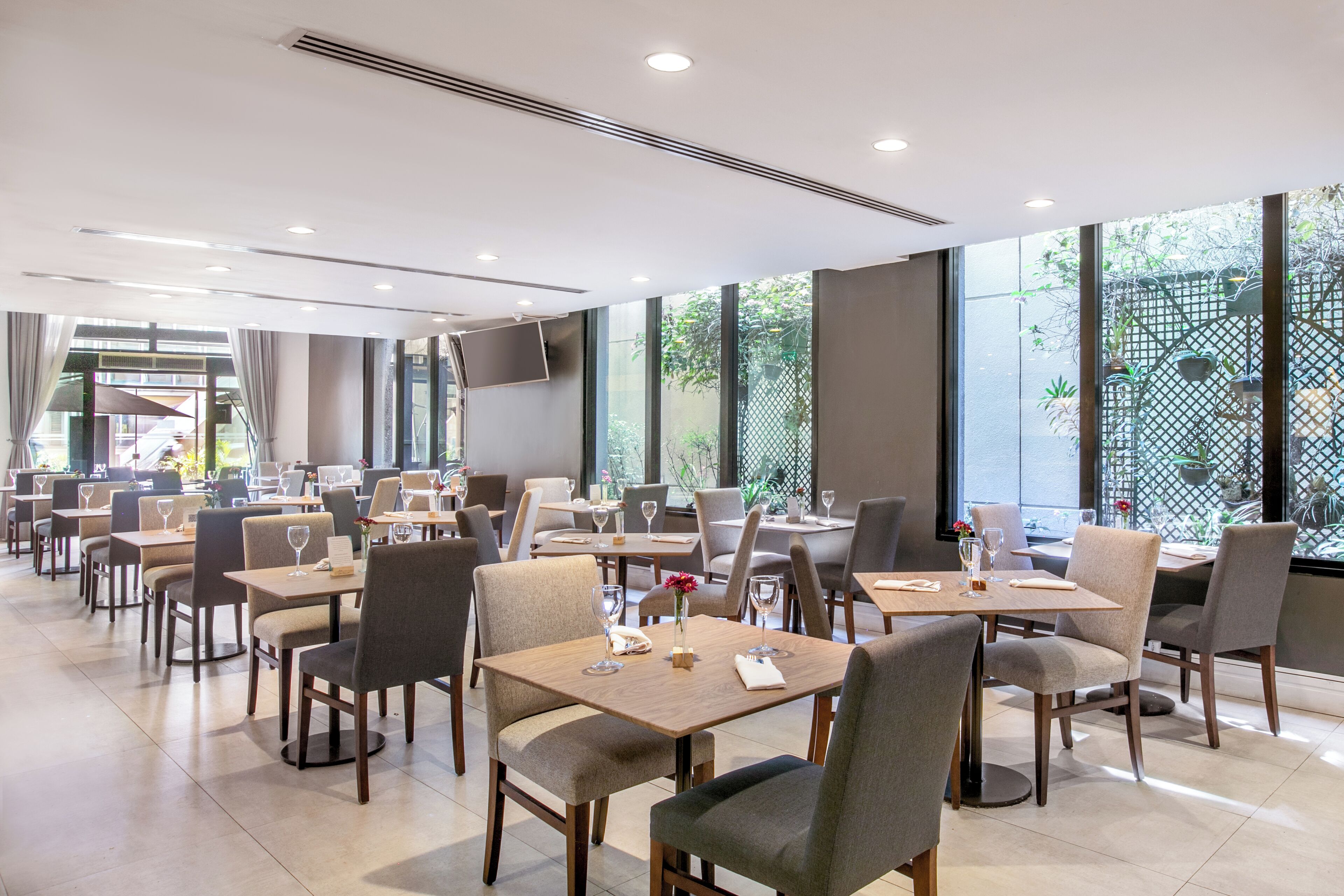 A modern restaurant with multiple tables and chairs, large windows, and a view of greenery outside.