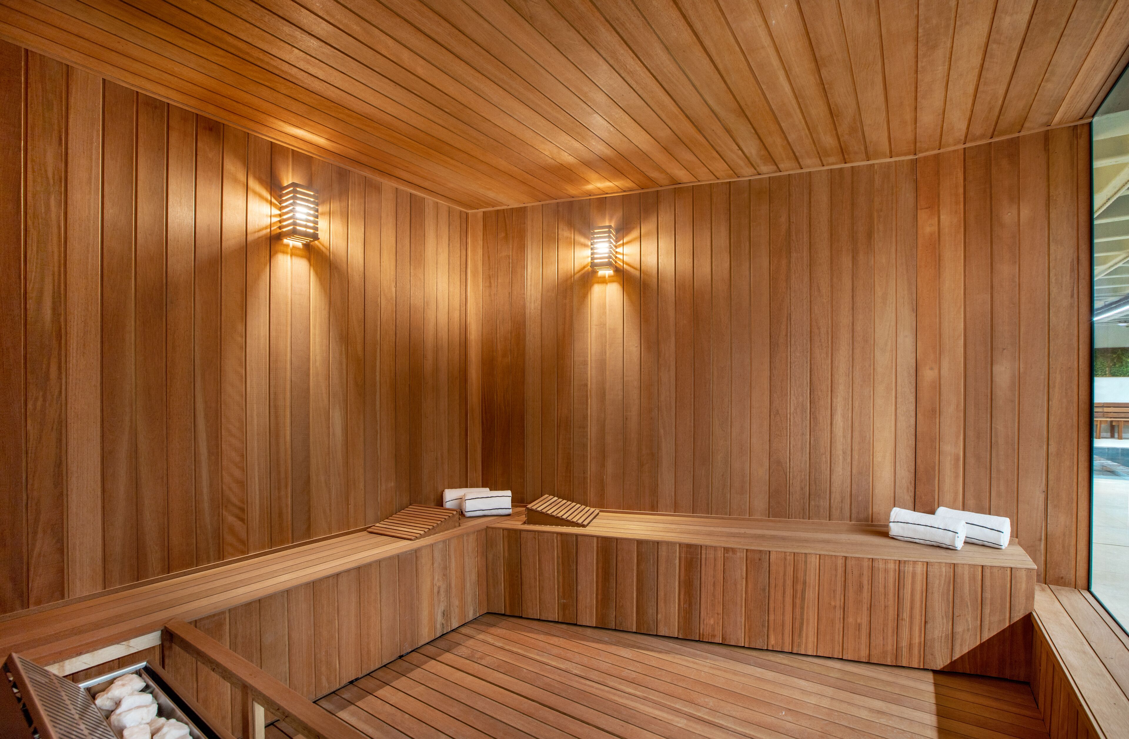 A sauna with wooden walls and benches, two wall-mounted light fixtures, and a view of the outside.