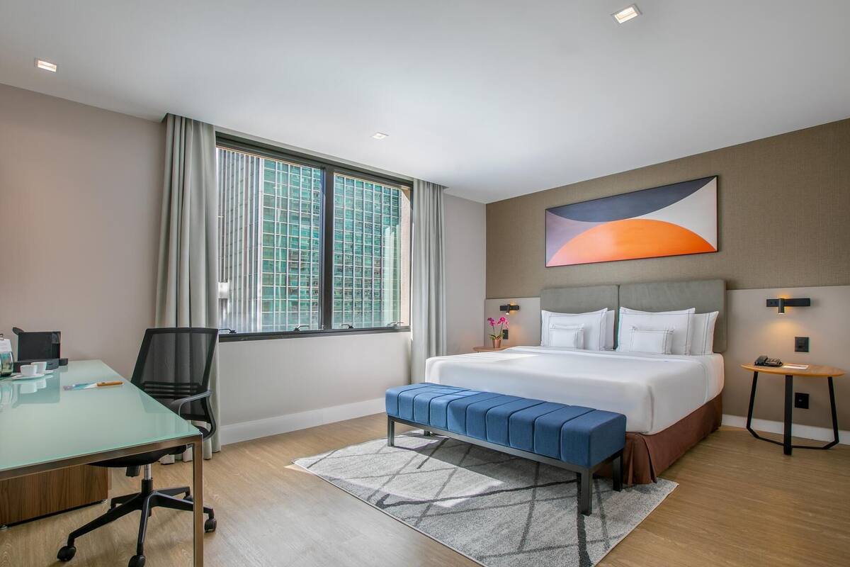 Habitación de hotel con cama, escritorio, silla, banco, alfombra y un cuadro en la pared.