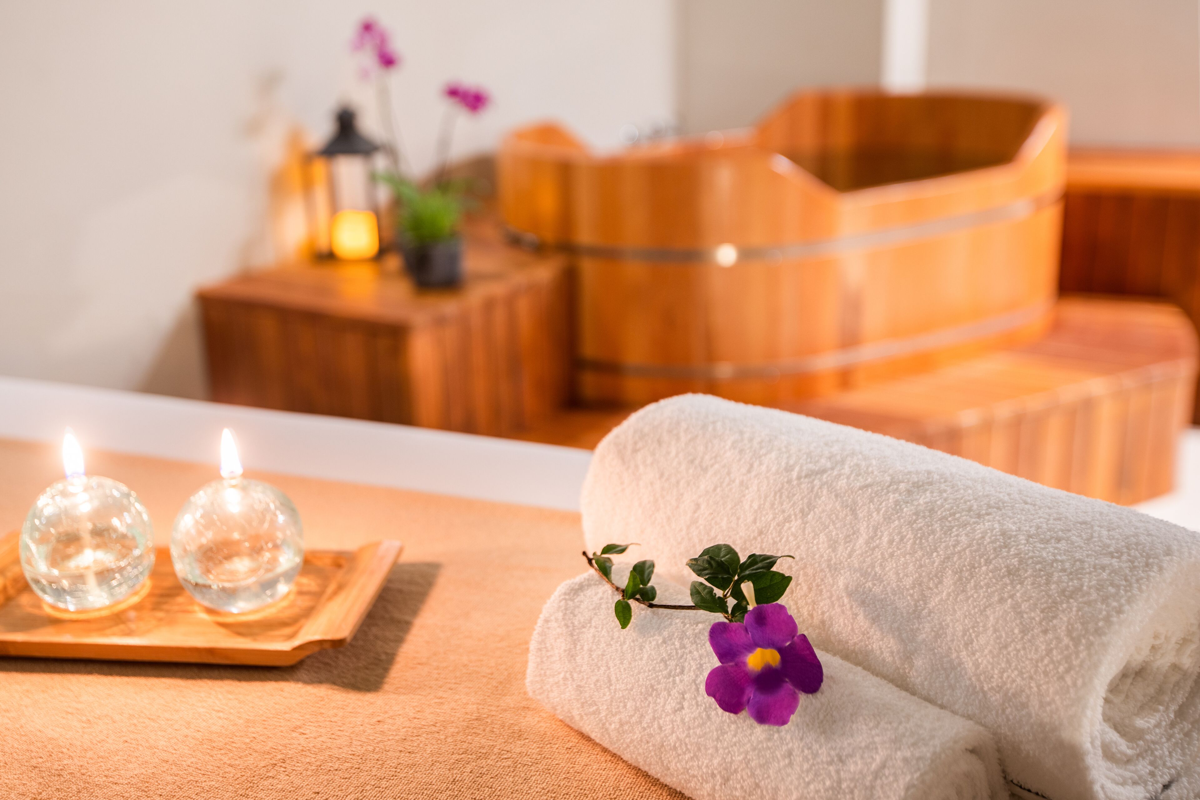 A spa setting with a wooden tub, a rolled white towel, and two lit candles on a wooden tray.
