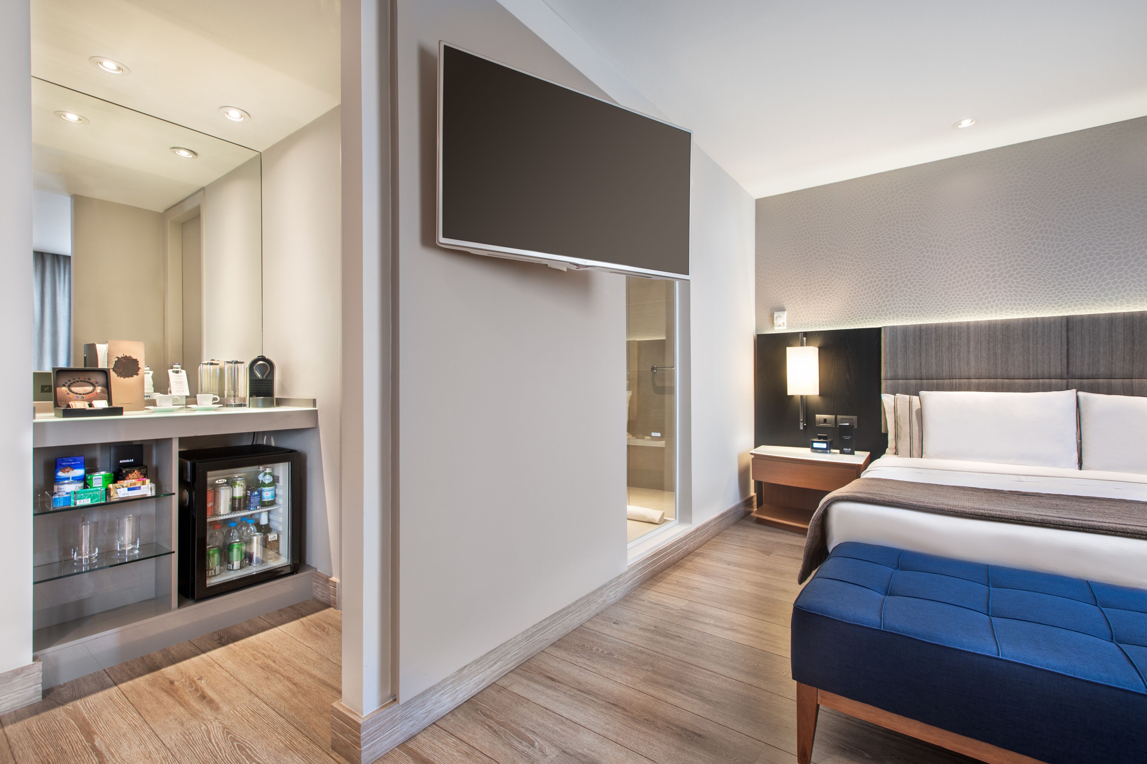 A modern hotel room with a large bed, a flat-screen TV mounted on the wall, a minibar, and a wooden floor.
