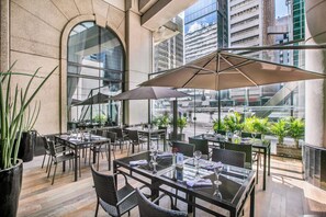 Ein Essbereich im Freien mit Tischen und Stühlen, großen Sonnenschirmen und Pflanzen in Töpfen.
