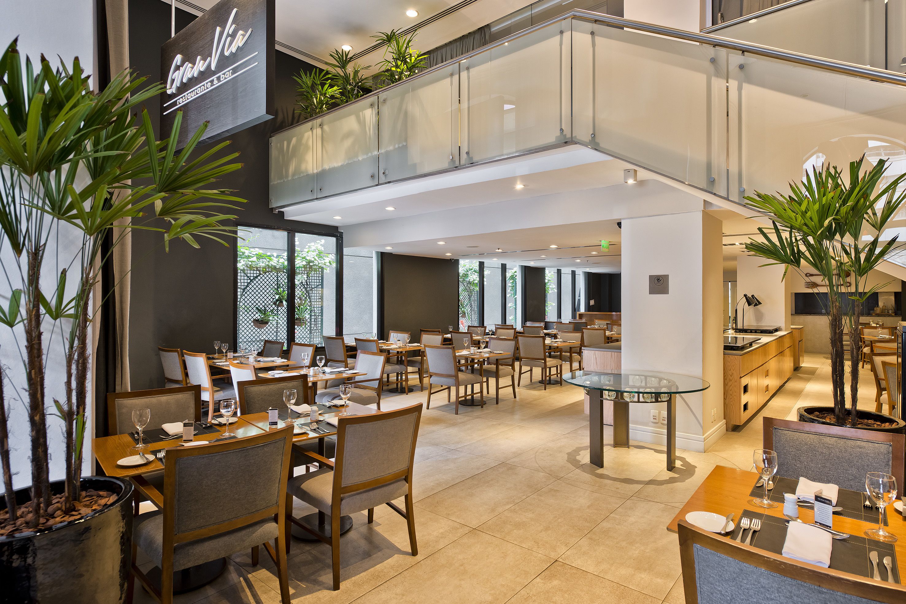 Un restaurante moderno con varias zonas de comedor, barandilla de cristal y plantas en macetas.