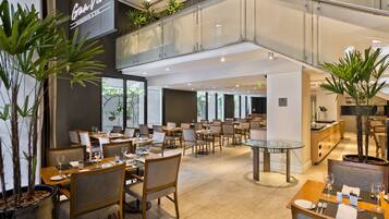 Un restaurante moderno con varias zonas de comedor, barandilla de cristal y plantas en macetas.