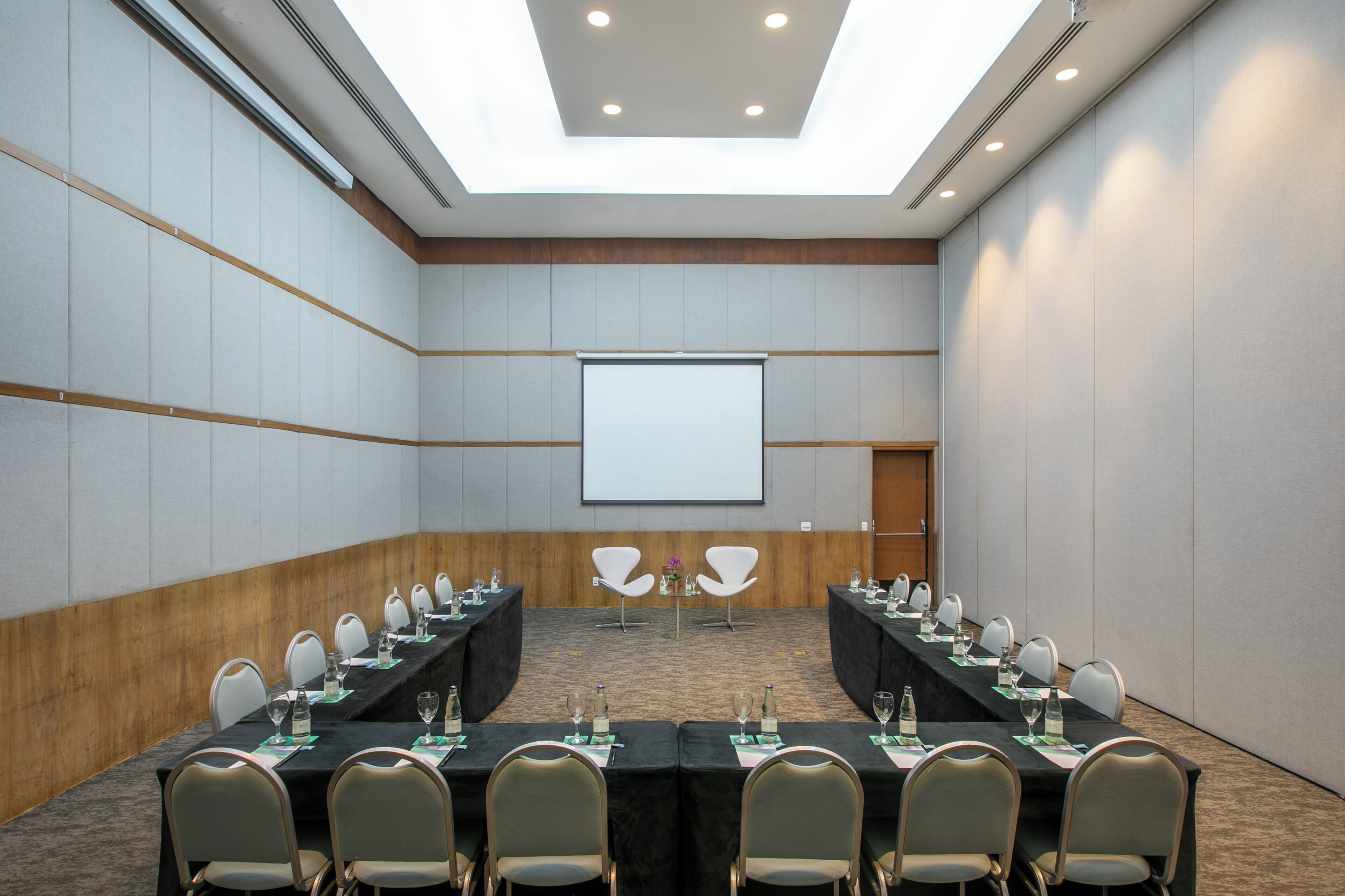 A conference room with a long table, chairs, a projector screen, and a wooden accent wall.