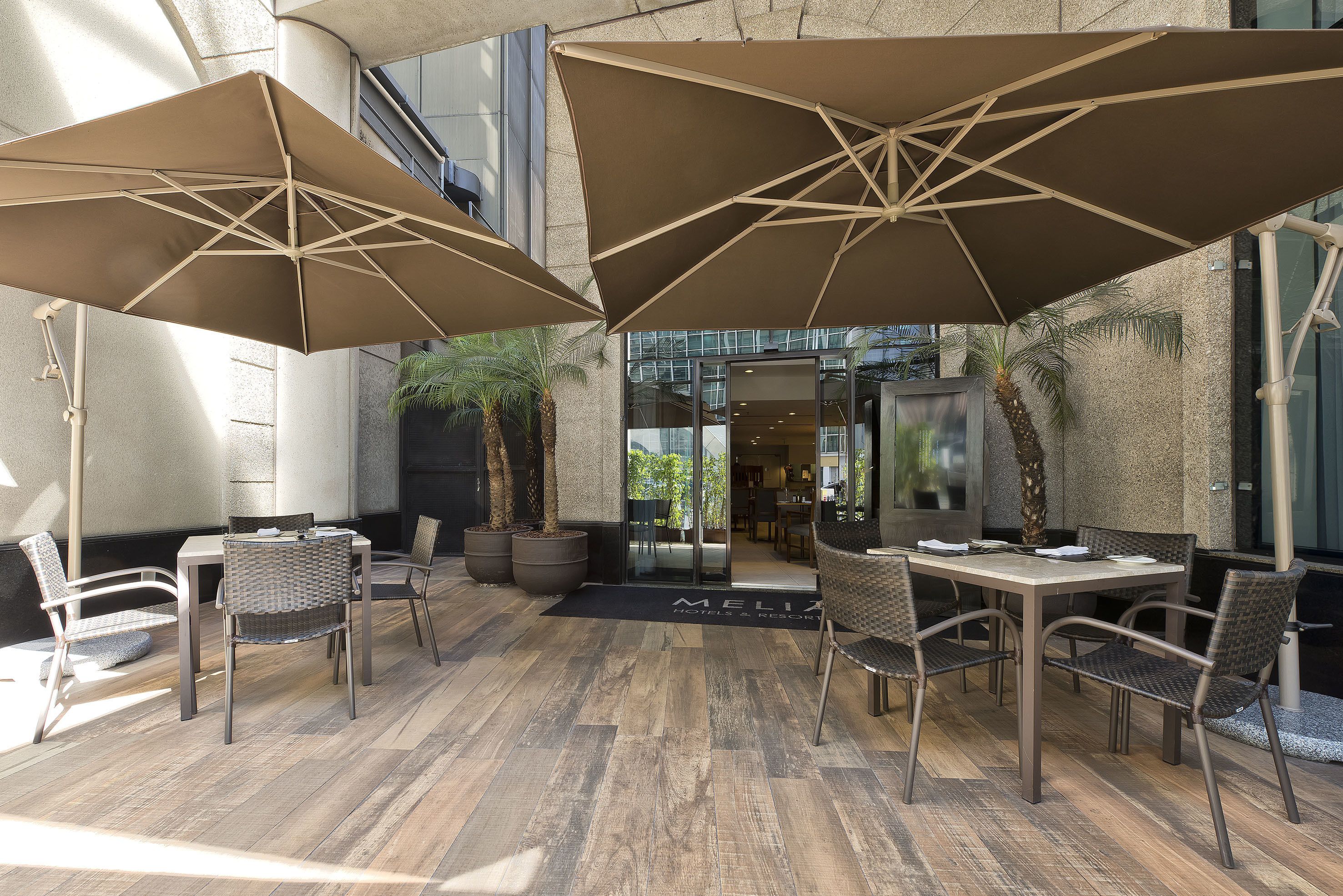 Eine Terrasse im Freien mit Tisch, Stühlen und großen Sonnenschirmen, sowie eine Tür mit der Aufschrift 'MELIA'.