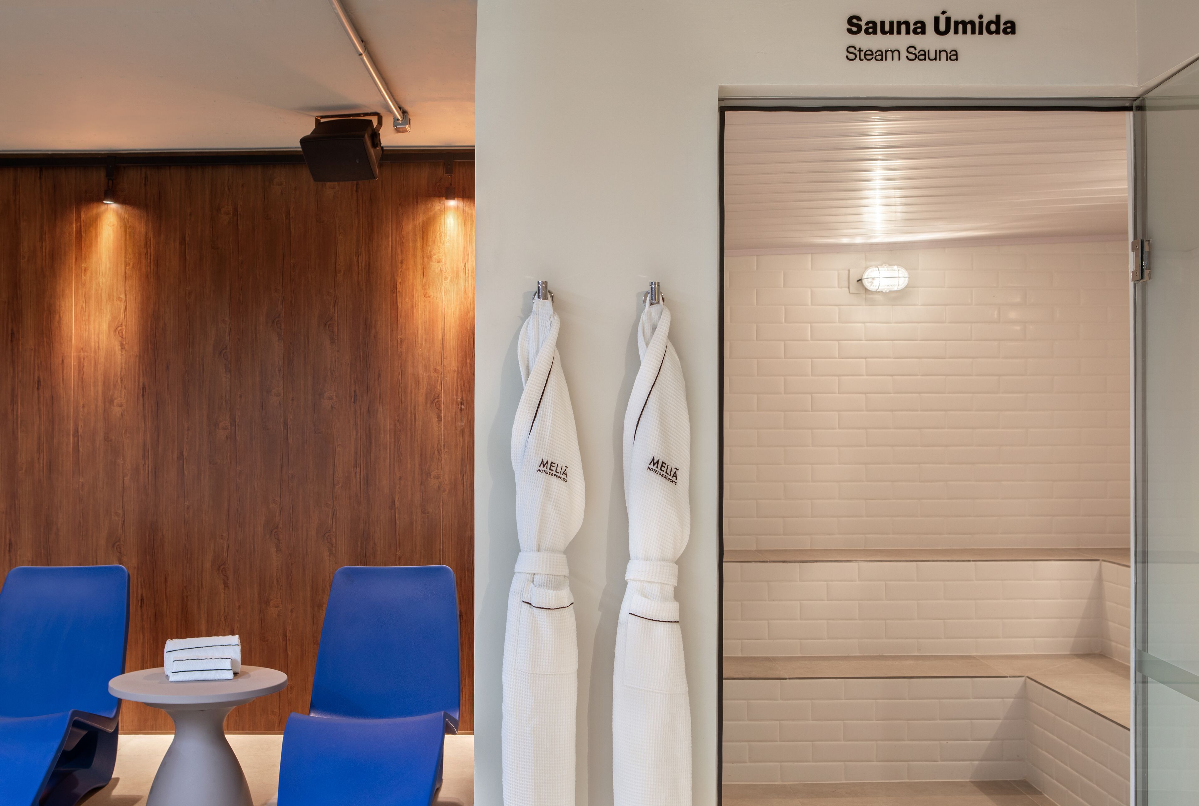 Uma sauna a vapor com paredes revestidas de azulejos brancos e uma parede de painéis de madeira ao fundo. Há duas toalhas brancas penduradas em ganchos e uma pequena mesa com uma pilha de livros.