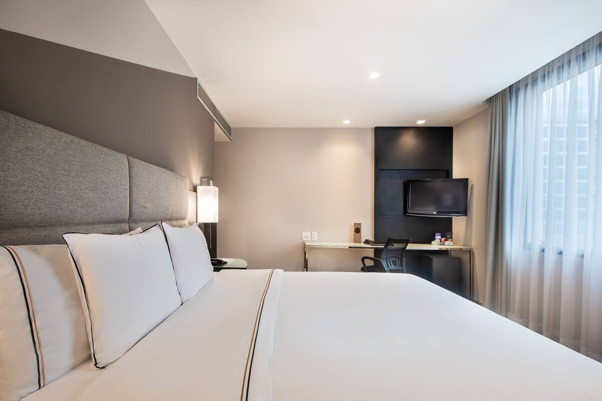Habitación de hotel con una cama grande, un escritorio con una silla, un televisor de pantalla plana montado en la pared y una ventana con cortinas.