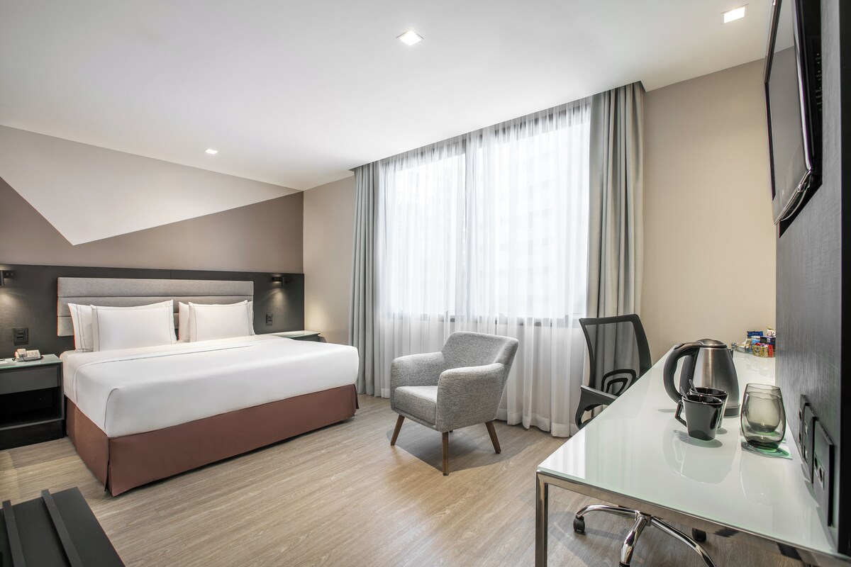 Una habitación de hotel moderna con una cama grande, un espacio de descanso con una silla, una mesa de centro y un televisor montado en la pared.