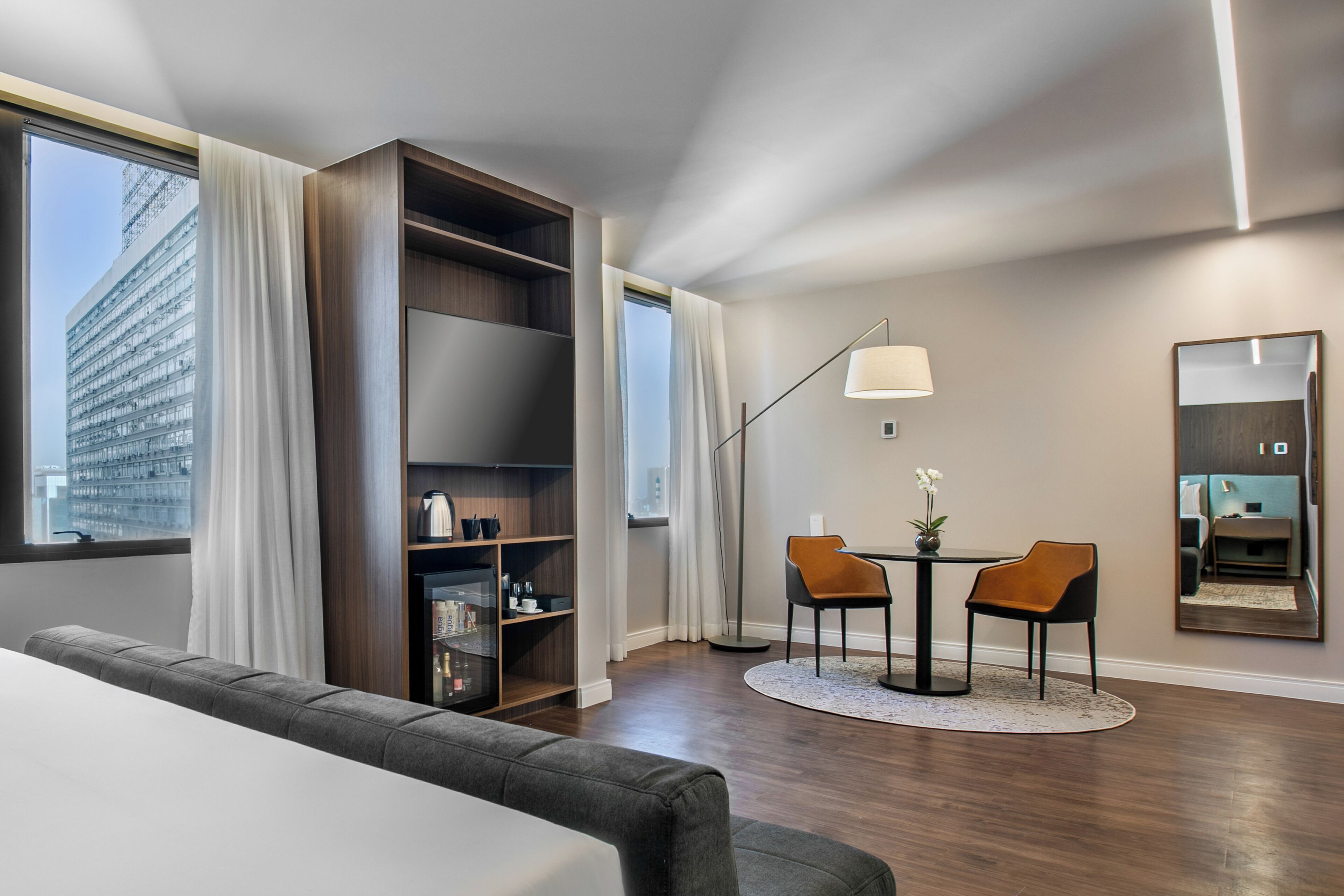 A modern hotel room with a large window, a built-in bookshelf, a round dining table with two chairs, a floor lamp, and a mirror on the wall.