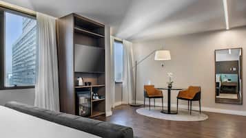 A modern hotel room with a large window, a built-in bookshelf, a round dining table with two chairs, a floor lamp, and a mirror on the wall.