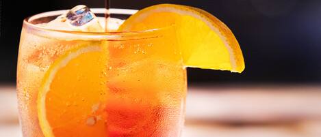 A glass of cocktail with an orange slice garnish and ice cubes, placed on a wooden coaster.