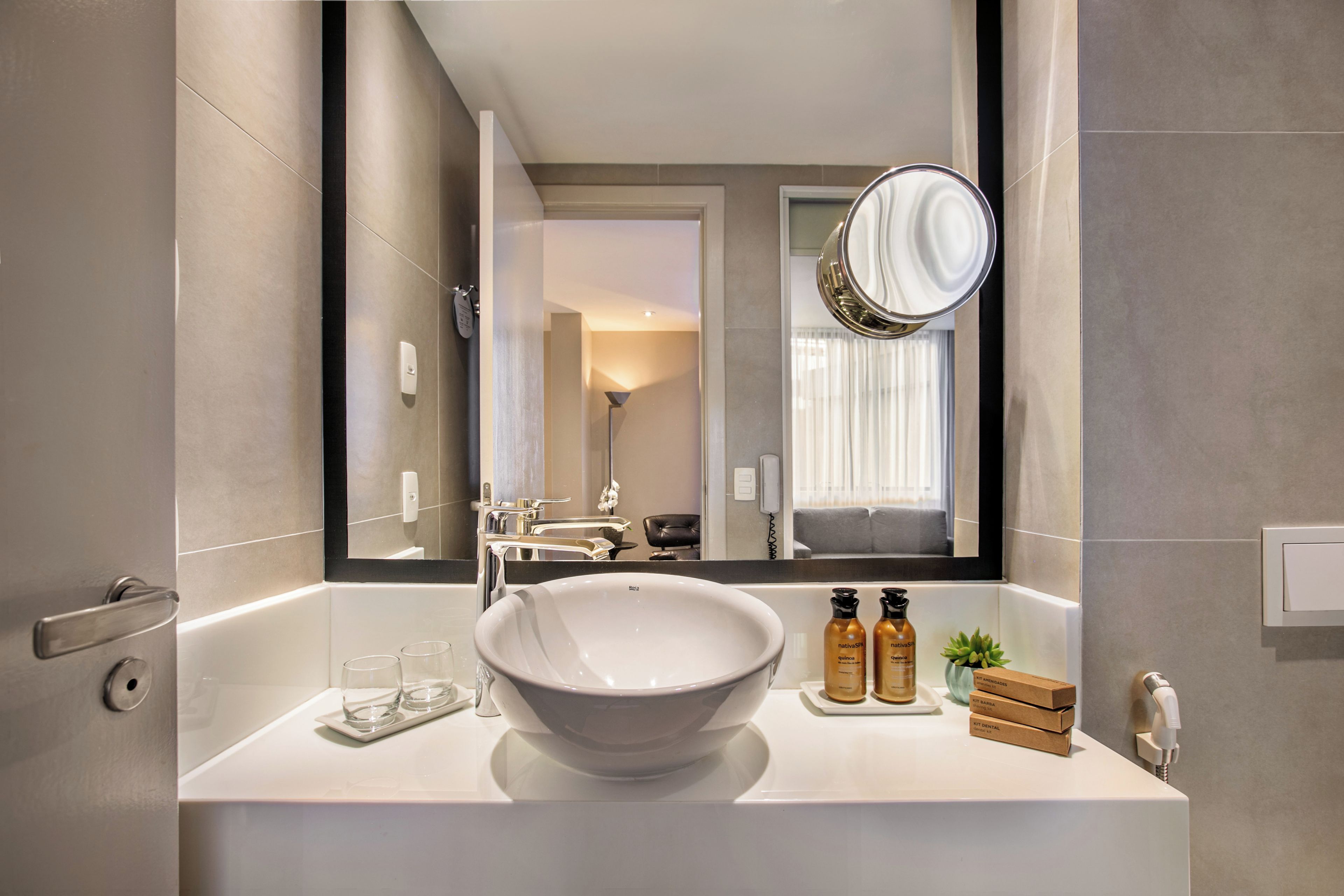 A modern bathroom with a large bathtub, a round mirror, and a vanity with a bowl sink. There are toiletries on the vanity and a potted plant beside the sink.